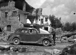 Un acompanyant de Francesc Josep de Liechtenstein revisant el motor d'un cotxe davant d'una casa molt danyada pels bombardeigs, al front d'Aragó